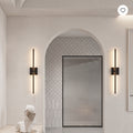 A modern bathroom interior with two slim black wall sconces mounted on cream walls on either side of an arched doorway. The sconces have vertical light bars that glow warm white.
