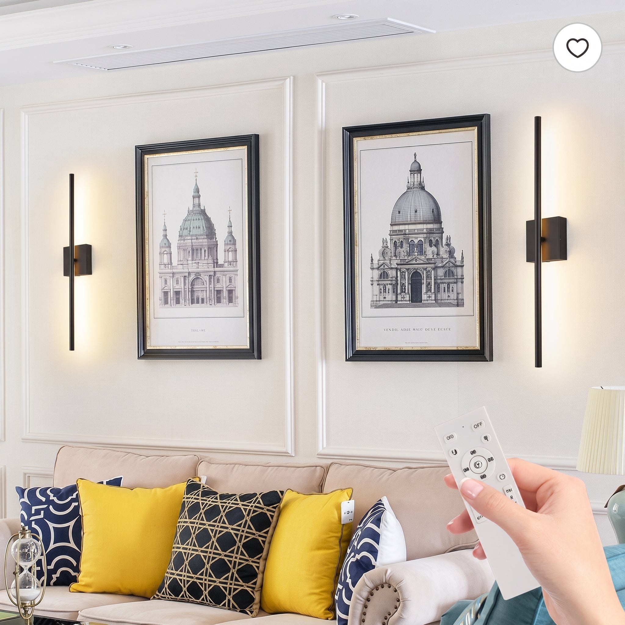 A lifestyle scene showing a hand holding a white remote control in front of a beige sofa with yellow and navy patterned pillows. Above the sofa, two black-framed architectural prints hang on a white paneled wall, flanked by two modern vertical wall sconces emitting warm light.