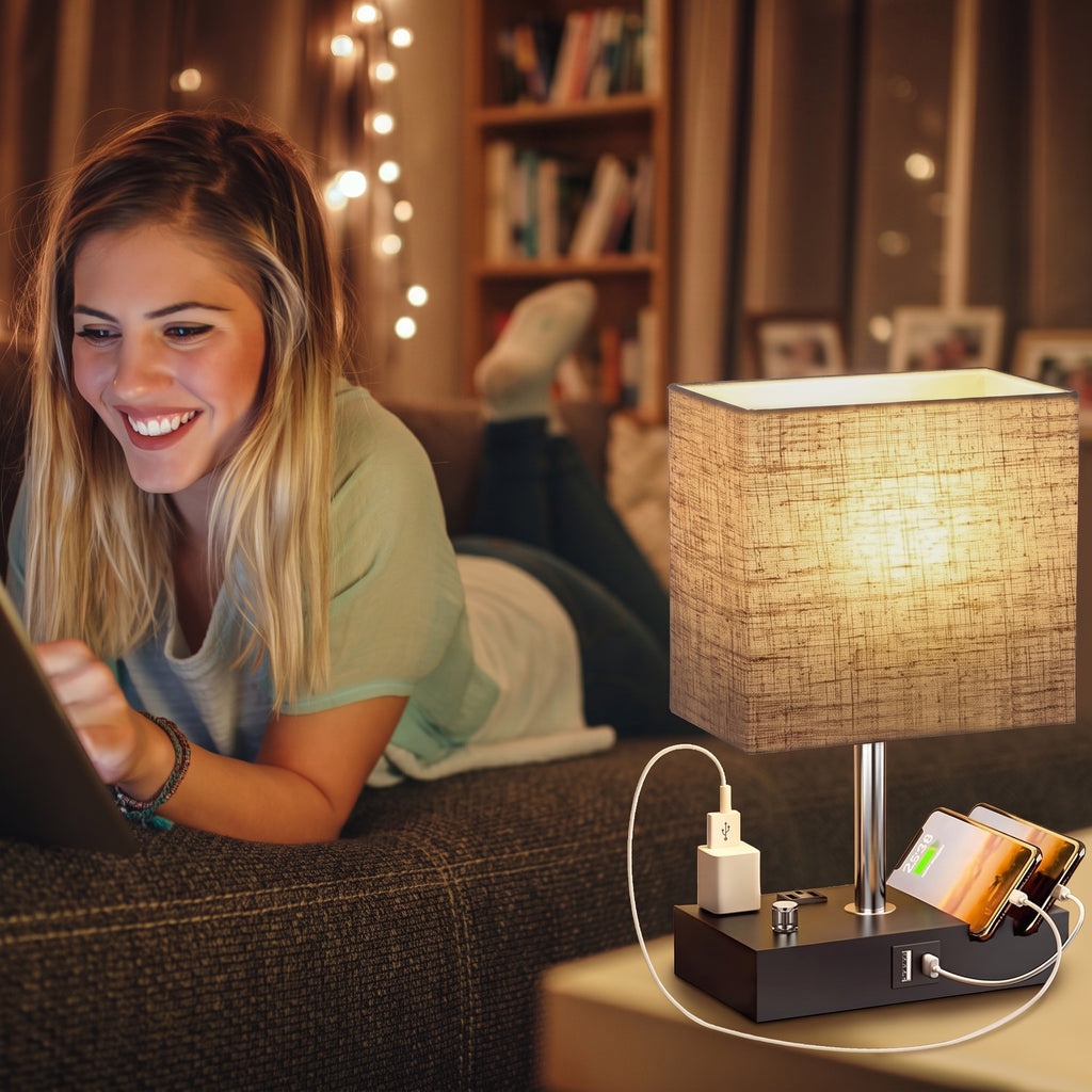 A woman lies on a couch in a cozy living room while using a laptop. In the foreground, a lit table lamp with a textured beige shade sits on a side table, charging a smartphone and other devices via built-in ports on its black base.