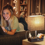 A woman lies on a couch in a cozy living room while using a laptop. In the foreground, a lit table lamp with a textured beige shade sits on a side table, charging a smartphone and other devices via built-in ports on its black base.
