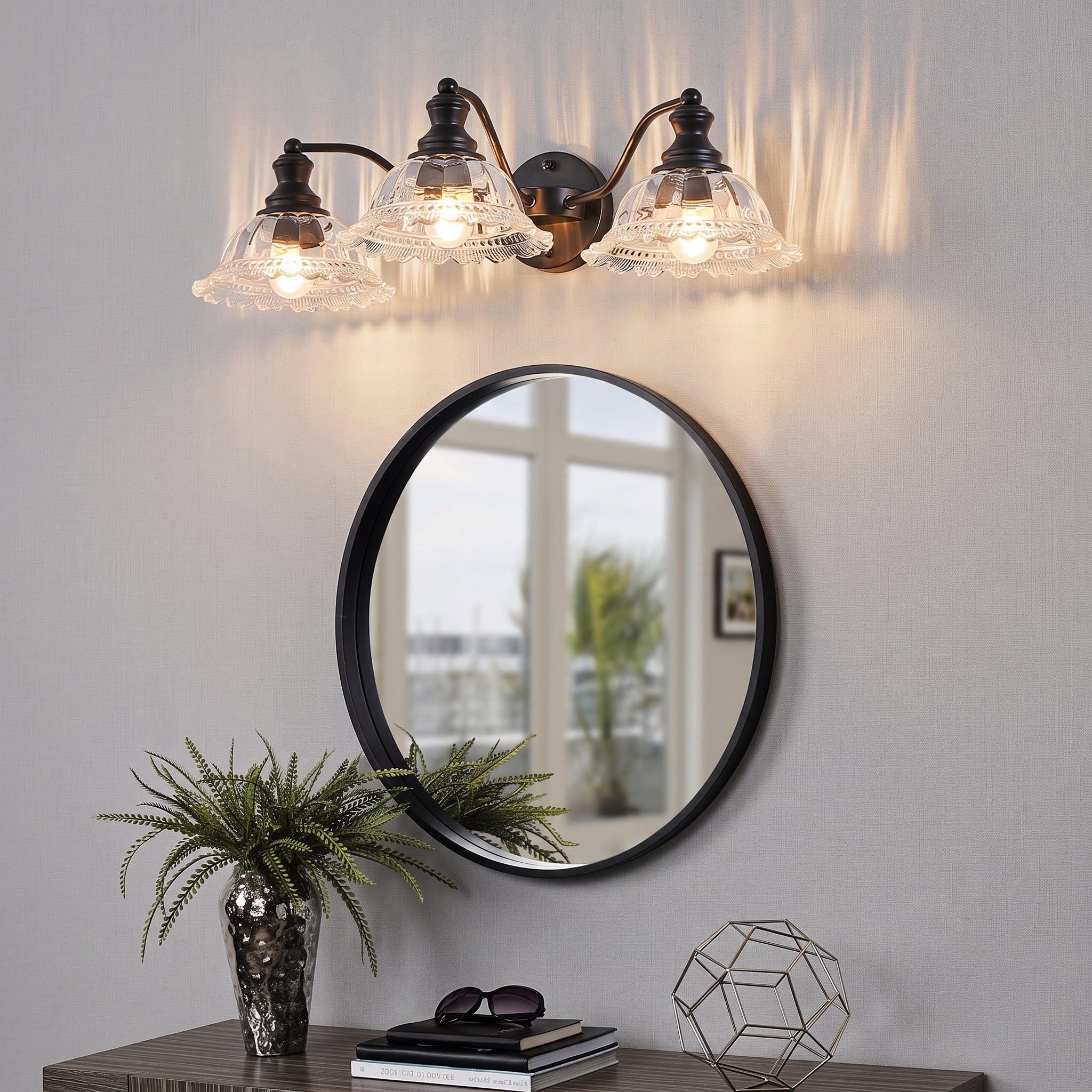 A three-light wall sconce with clear textured glass shades and an antique bronze finish is mounted above a round black-framed mirror on a light gray wall. A console table below holds a potted fern, books, and a geometric decor object.
