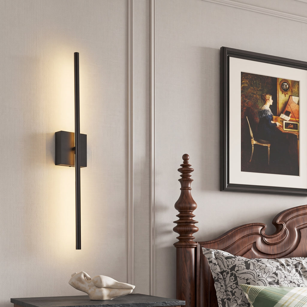 A modern vertical linear wall sconce with a matte black finish and warm LED lighting, mounted on a light gray paneled wall in a bedroom setting next to a wooden bed frame and framed artwork.