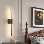 A modern vertical linear wall sconce with a matte black finish and warm LED lighting, mounted on a light gray paneled wall in a bedroom setting next to a wooden bed frame and framed artwork.