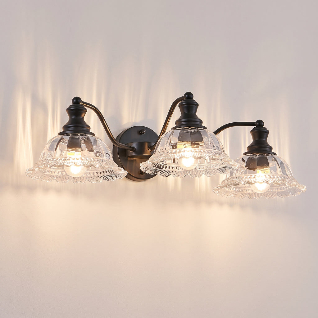 A three-light wall sconce with a dark metal mounting plate and curved arms, featuring clear ribbed glass shades that glow with warm light against a plain wall.