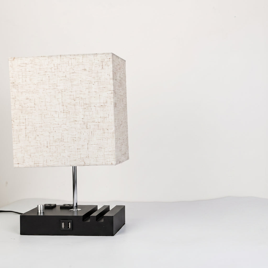 A modern table lamp with a square textured off-white fabric shade, a slim silver metal stem, and a black rectangular base featuring a USB port and decorative side slots, set against a plain white background.