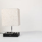 A modern table lamp with a square textured off-white fabric shade, a slim silver metal stem, and a black rectangular base featuring a USB port and decorative side slots, set against a plain white background.