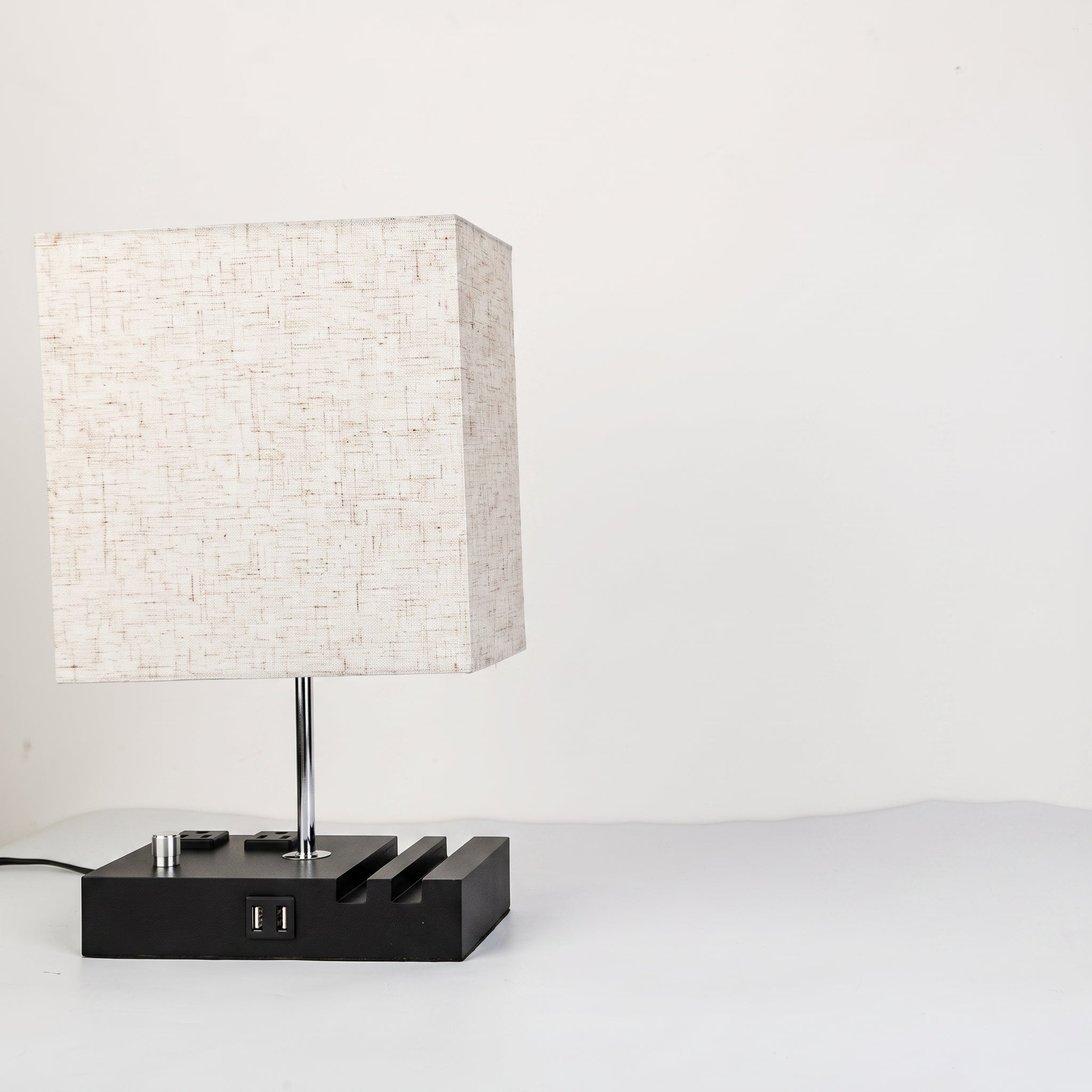 A modern table lamp with a square textured off-white fabric shade, a slim silver metal stem, and a black rectangular base featuring a USB port and decorative side slots, set against a plain white background.