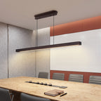 A modern linear LED pendant light in a matte black finish hangs over a wooden conference table in a contemporary office meeting room with gray chairs and a terracotta and white accent wall.