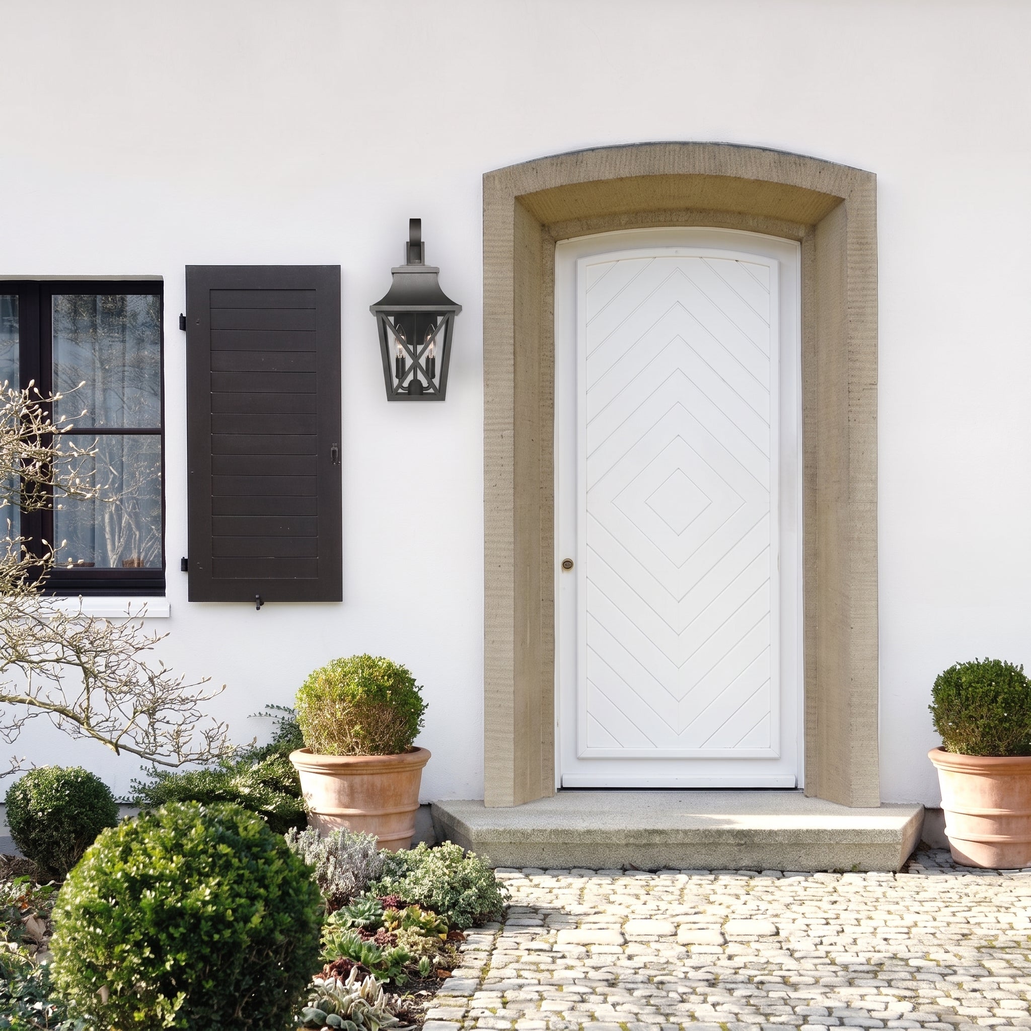 A white front door with a chevron panel design, set in a textured beige stone archway. The door is mounted on a white stucco wall next to a black shuttered window and a black wall lantern.