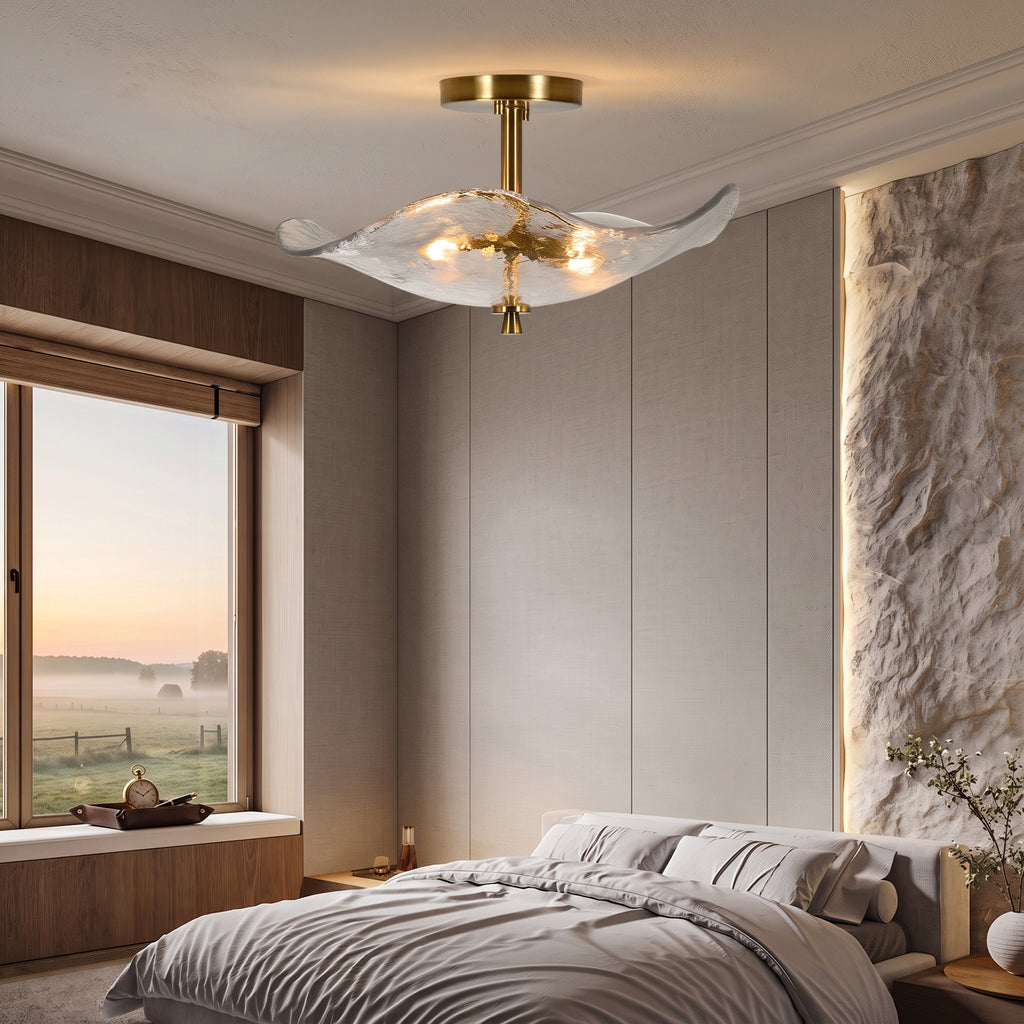 A modern ceiling light fixture with a gold-toned brass frame and a translucent, petal-like glass shade hangs over a bed in a contemporary bedroom with warm ambient lighting.