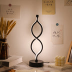 A modern black spiral LED table lamp illuminates a styled desk with books, wheat stalks, and a bubbly candle. Its unique, flowing design provides a soft glow. Text: 疏宗 青味至今 遠可請他, 吟味不是 一道而旬, We are designed to unleash cultivation, 摧花 狀果, Good luck, thank you, come on, yeah.