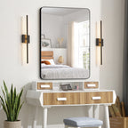 A large rectangular wall mirror with a thin black frame hangs above a white vanity table with wood-grain drawer fronts. The scene is styled in a bright bedroom, with the mirror reflecting a bed and window.