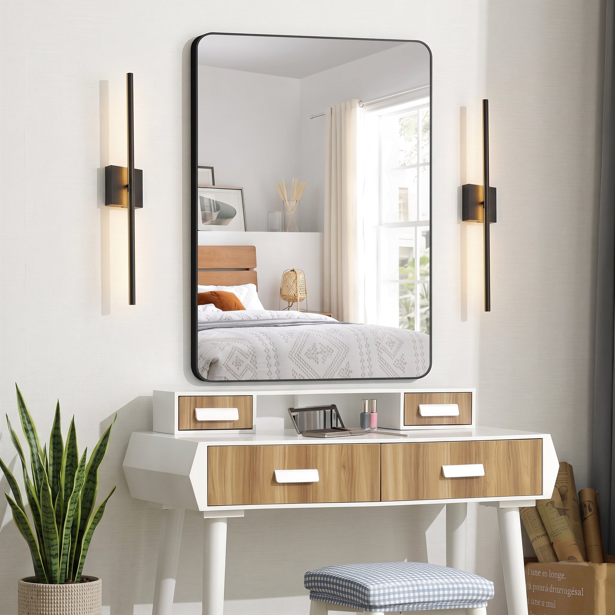 A large rectangular wall mirror with a thin black frame hangs above a white vanity table with wood-grain drawer fronts. The scene is styled in a bright bedroom, with the mirror reflecting a bed and window.