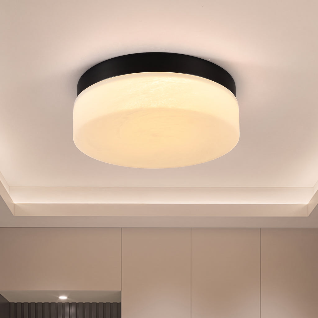 A modern flush-mount ceiling light with a black circular frame and a frosted white glass diffuser, emitting a warm yellow light. The light is mounted on a white ceiling with a recessed tray.