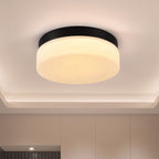 A modern flush-mount ceiling light with a black circular frame and a frosted white glass diffuser, emitting a warm yellow light. The light is mounted on a white ceiling with a recessed tray.