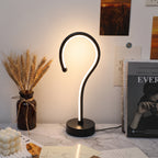 A modern curved LED table lamp with a matte black finish and a glowing white light, placed on a marble-patterned surface in a styled interior setting with books, a vase of wheat, and wall art in the background.