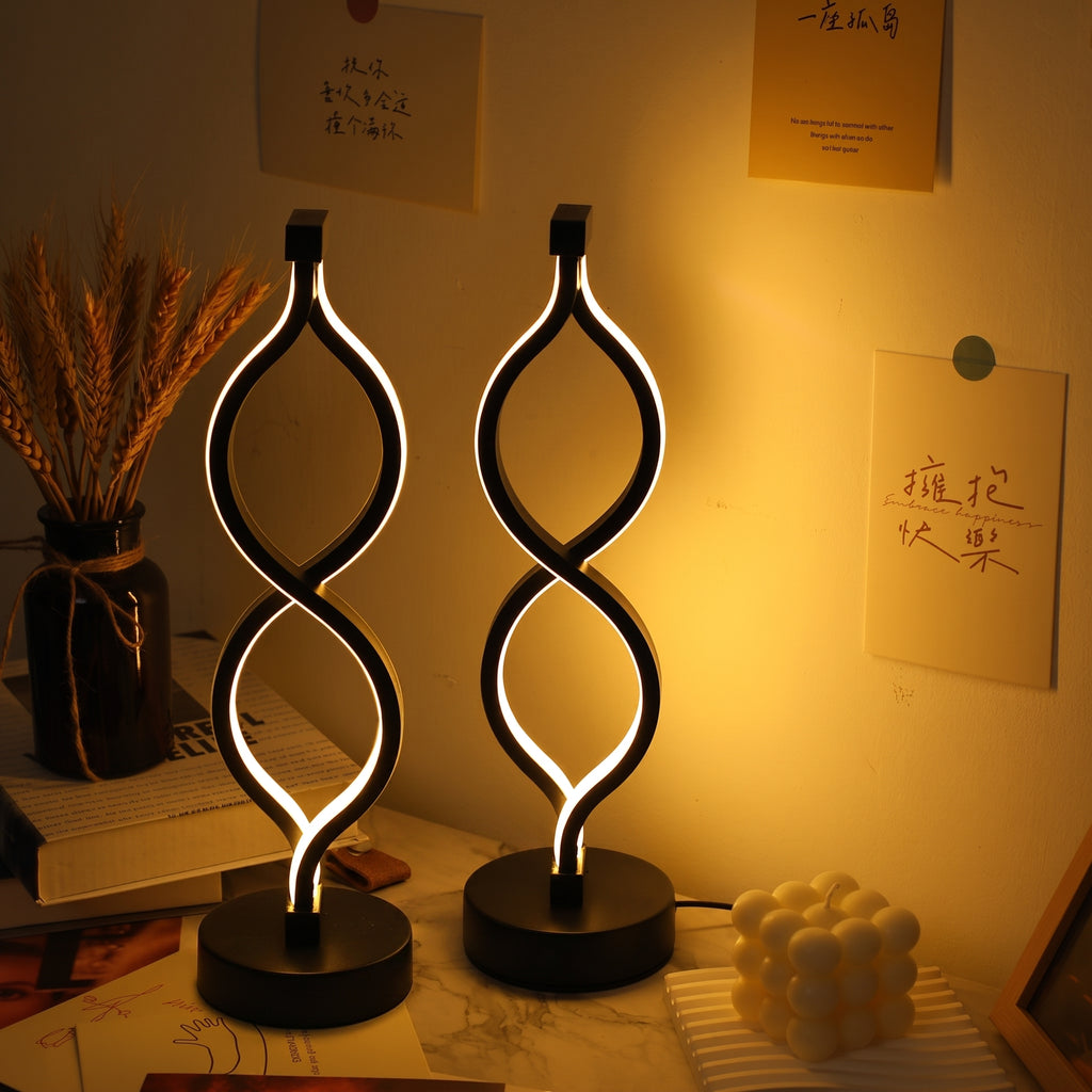 Two modern twisted-spiral LED table lamps with black bases, glowing with warm white light, set on a desk in a warmly lit room with handwritten notes on the wall.