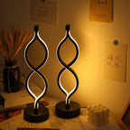 Two modern twisted-spiral LED table lamps with black bases, glowing with warm white light, set on a desk in a warmly lit room with handwritten notes on the wall.