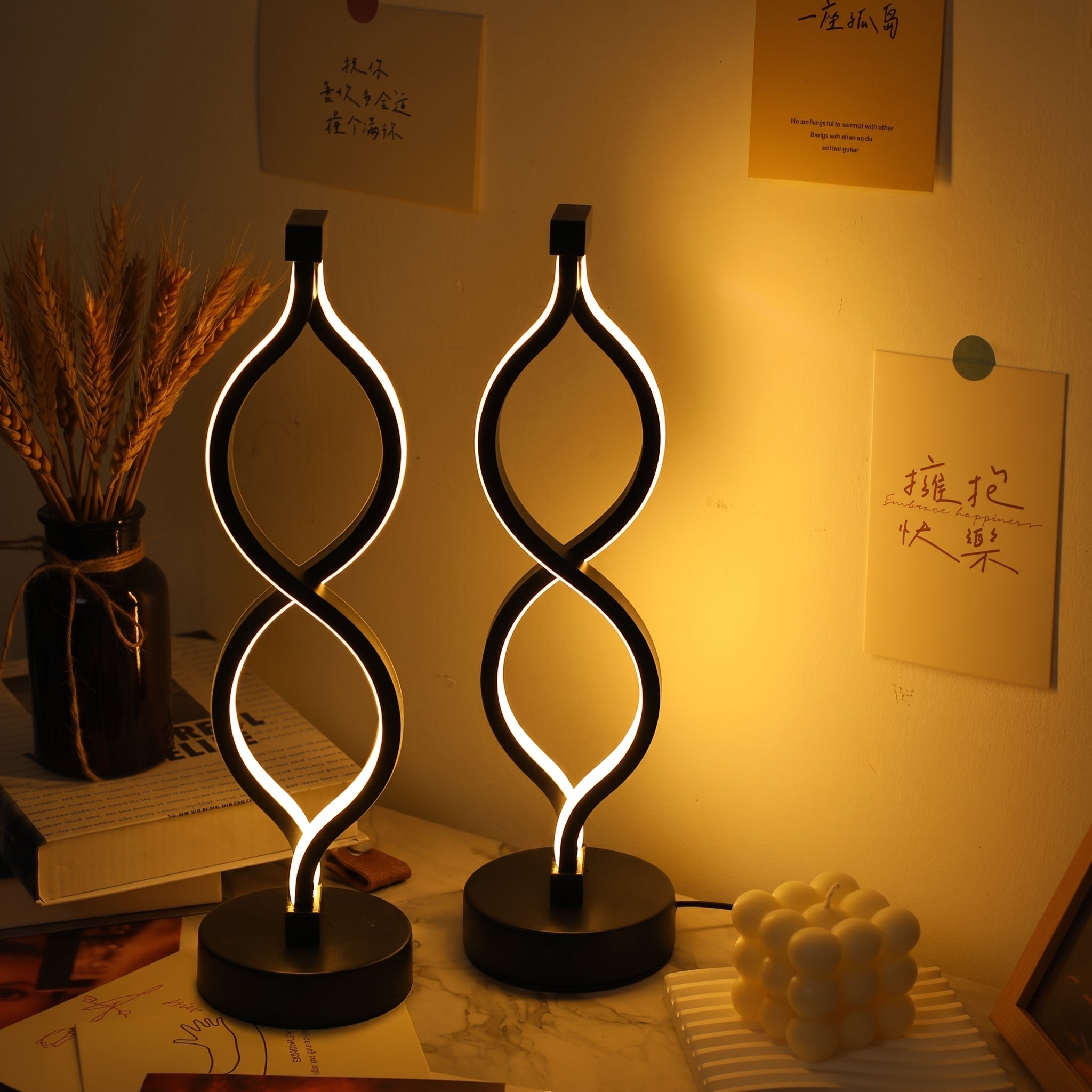 Two modern twisted-spiral LED table lamps with black bases, glowing with warm white light, set on a desk in a warmly lit room with handwritten notes on the wall.
