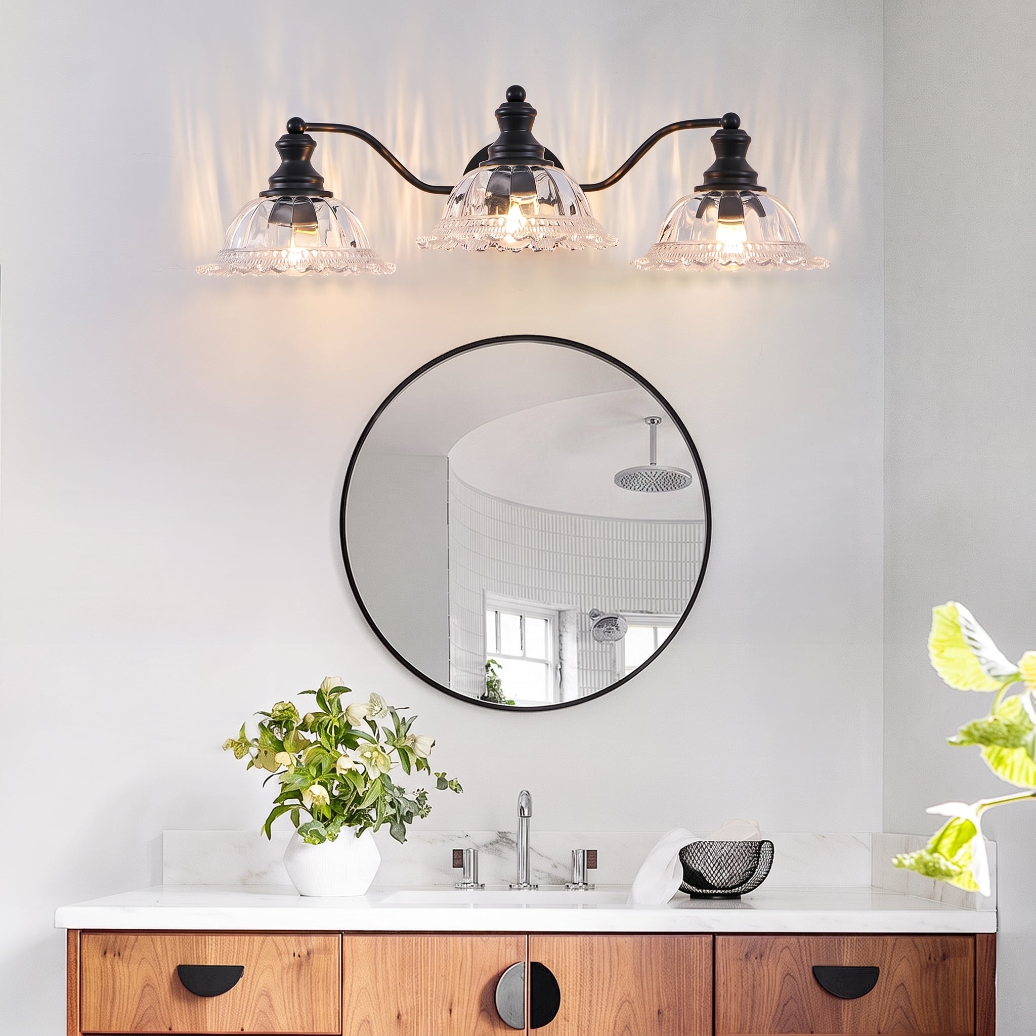 A three-light wall sconce with black metal arms and clear glass shades is mounted above a round black-framed mirror in a bright bathroom setting.