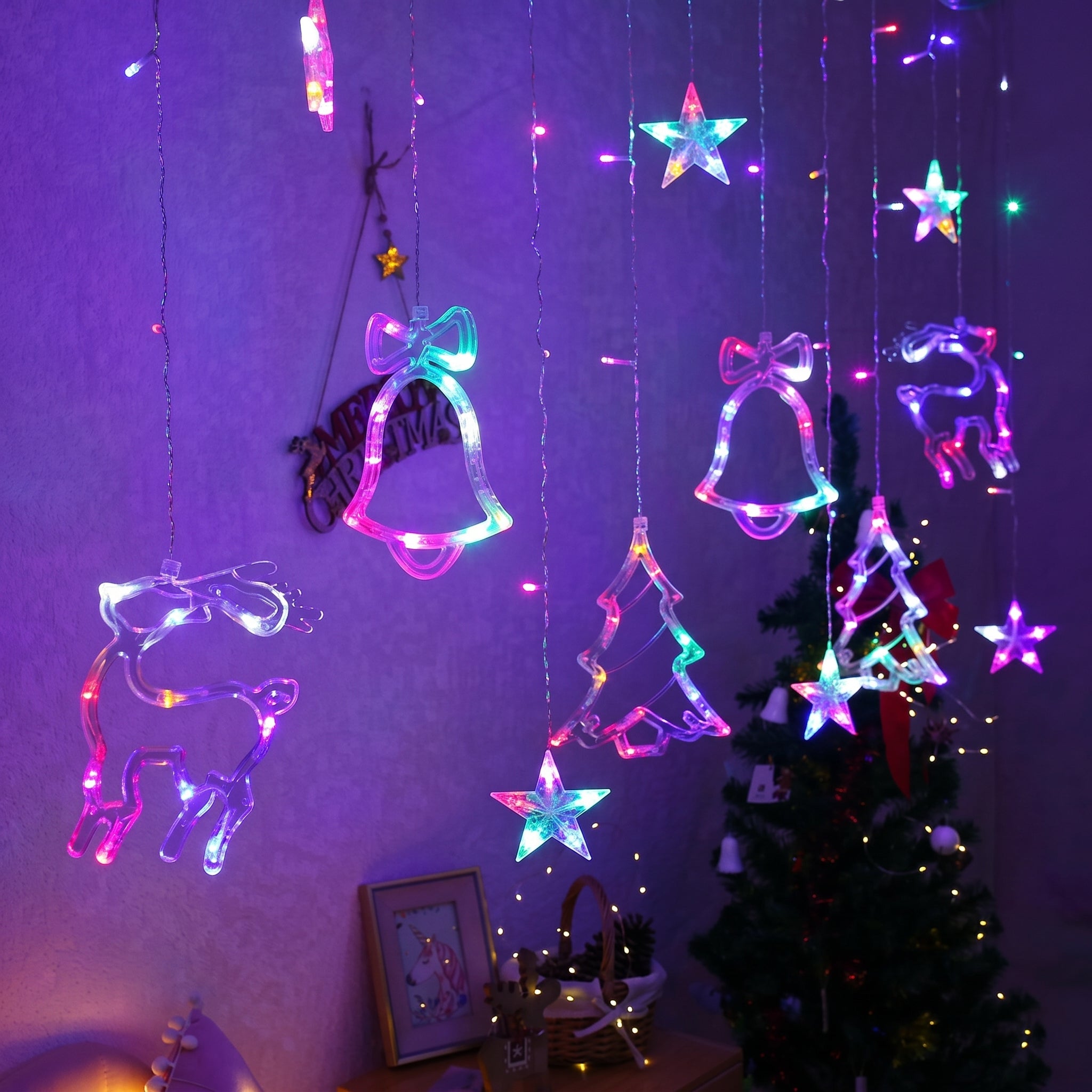 Multicolor LED hanging Christmas string lights shaped like stars, bells, bows, and a reindeer, displayed on a wall above a decorated Christmas tree.