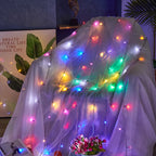 A sheer white mesh fabric draped over a chair, decorated with multicolored fairy string lights in red, green, blue, yellow, and pink. The background shows a dark blue wall, a decorative leaf print, and a small potted plant.