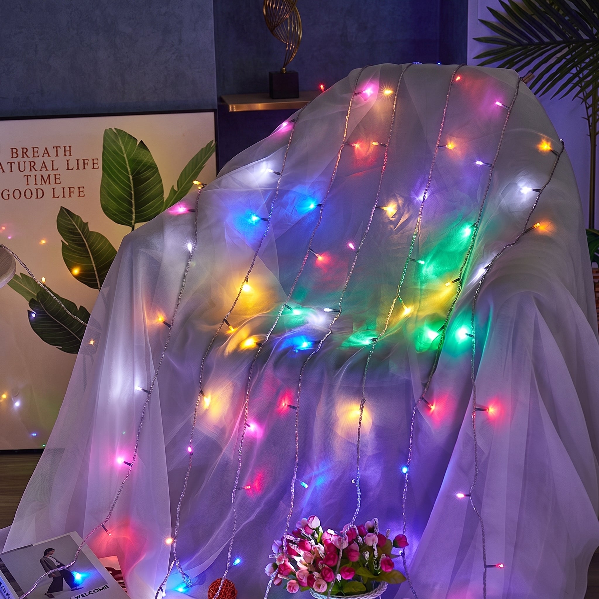 A sheer white mesh fabric draped over a chair, decorated with multicolored fairy string lights in red, green, blue, yellow, and pink. The background shows a dark blue wall, a decorative leaf print, and a small potted plant.
