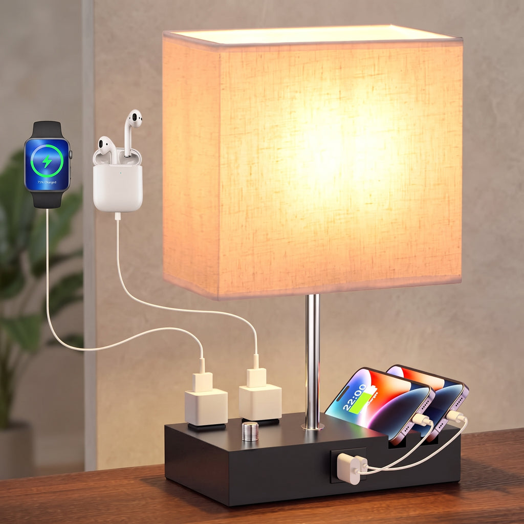 A modern table lamp with a glowing warm beige fabric shade, a slim silver metal stem, and a black rectangular charging base. The base includes multiple built-in charging ports and accessories: a smartwatch on the left, white earbuds in a charging case, two white power adapters, and two smartphones on the right.