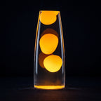 A clear glass lava lamp-style vase with three glowing orange-yellow blobs floating in transparent liquid, photographed against a black background.