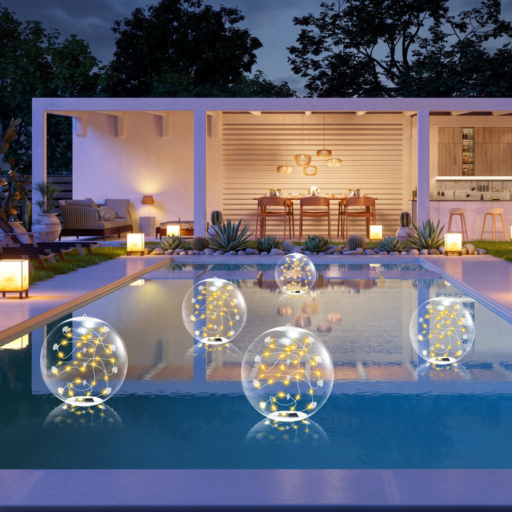 Five clear spherical floating light decorations with warm yellow string lights are arranged on the surface of a swimming pool at dusk, with a modern outdoor patio and lit dining area in the background.
