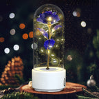 A preserved blue rose with glowing warm fairy lights inside a clear glass dome, set on a white cylindrical base. The product is photographed on a wooden surface with a blurred Christmas tree and ornaments in the background.
