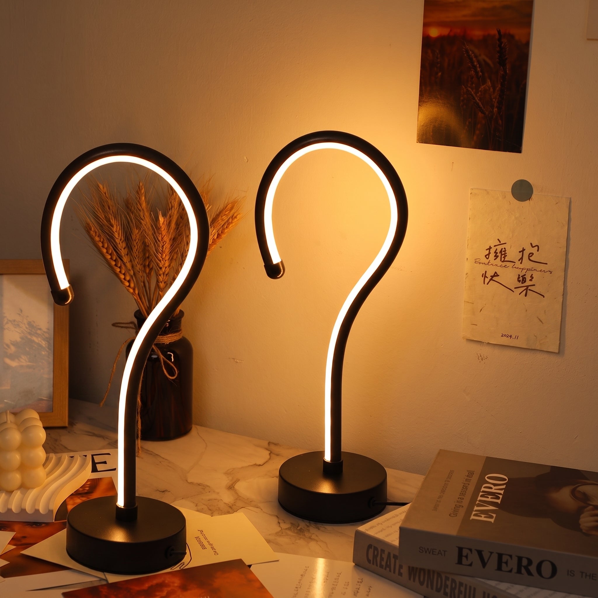Two modern curved LED table lamps with black bases and glowing white light tubes, placed on a marble-like surface in a warmly lit indoor setting with books, a vase of dried wheat, and wall art visible.