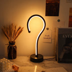 A modern curved LED table lamp with a black base and a glowing white light tube, styled on a marble desk next to a vase of wheat and books.