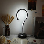 A modern black LED desk lamp with a curved, hook-like shape stands on a round base, casting a bright white light. It is styled on a marble tabletop next to a vase of dried wheat, an envelope with a red wax seal, and books against a softly lit wall.