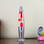 A silver and clear lava lamp with red wax blobs, standing on a wooden table against a beige wall, with a colorful toy partially visible on the right.