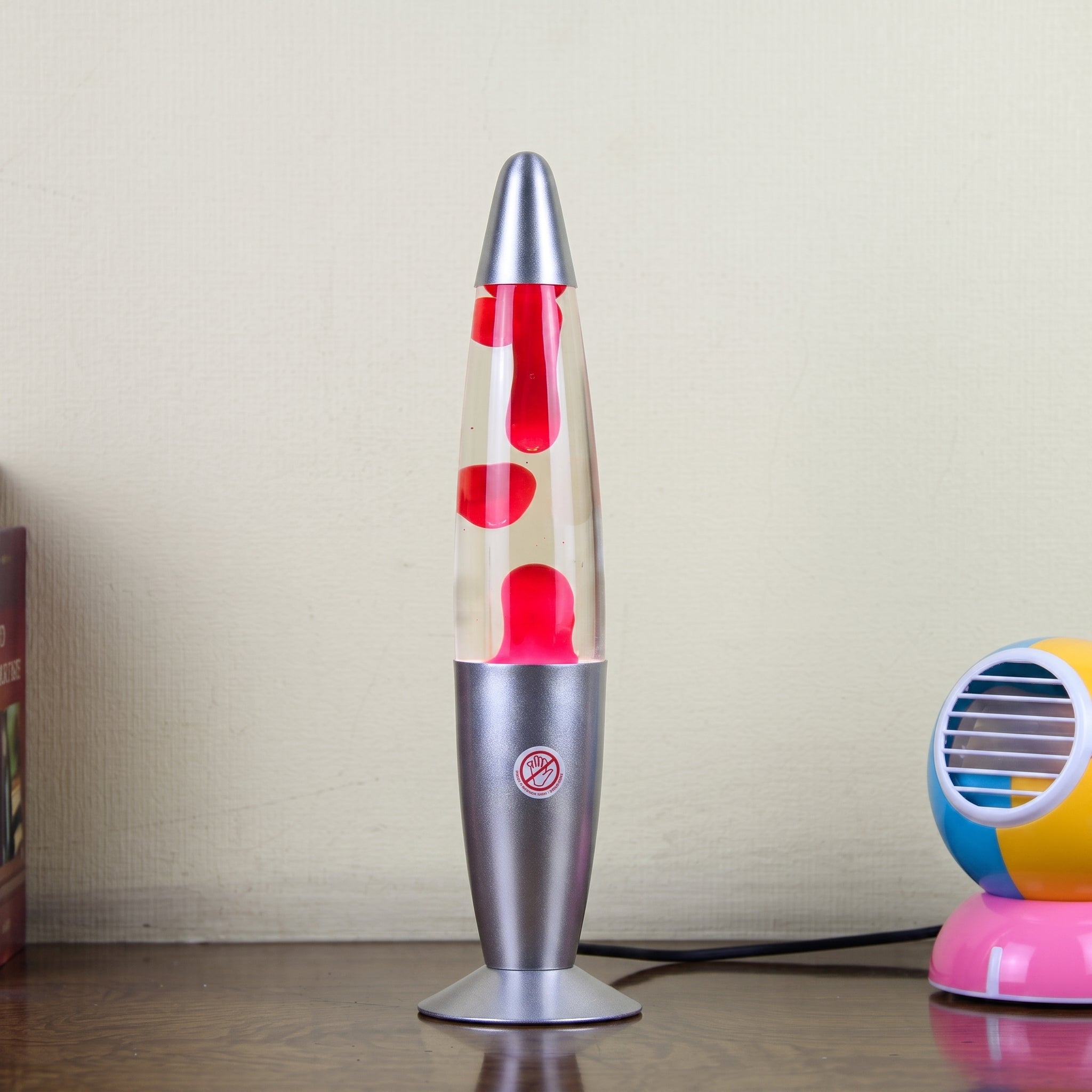 A silver and clear lava lamp with red wax blobs, standing on a wooden table against a beige wall, with a colorful toy partially visible on the right.