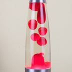 A classic lava lamp with a clear glass body, filled with bright red wax blobs and a silver metal base and top cap, set against a plain beige background.