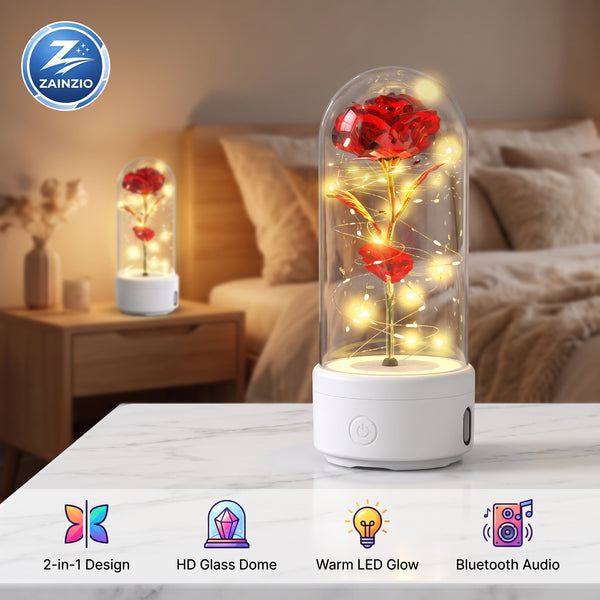A glass dome LED lighted rose night light on a white base, shown lit on a marble tabletop in a bedroom setting with a matching smaller version in the background. Features a ZAINZIO brand logo and feature icons along the bottom.