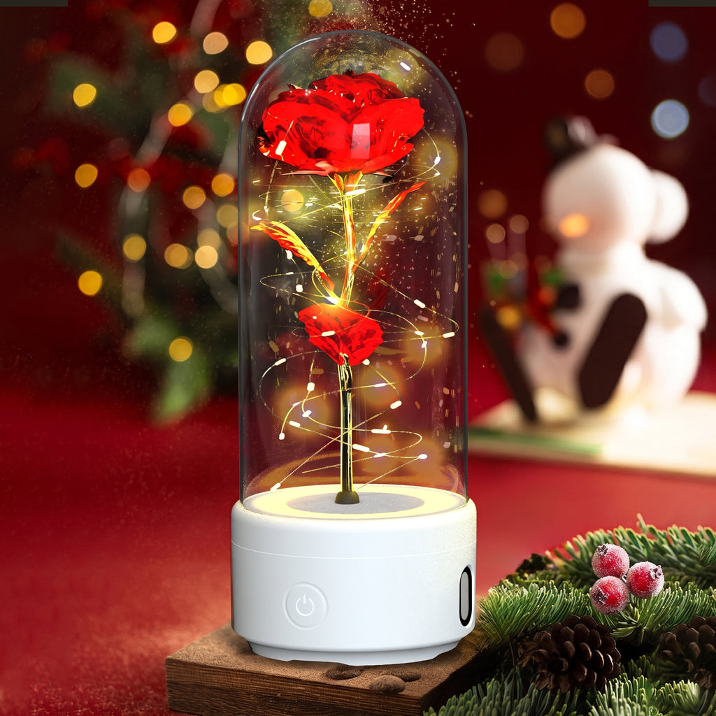 A decorative glass dome light holding two glowing red roses with warm fairy lights, set on a white cylindrical base. The scene is styled with a blurred Christmas tree and snowman in the background, on a red surface with pine branches and pinecones in the foreground.