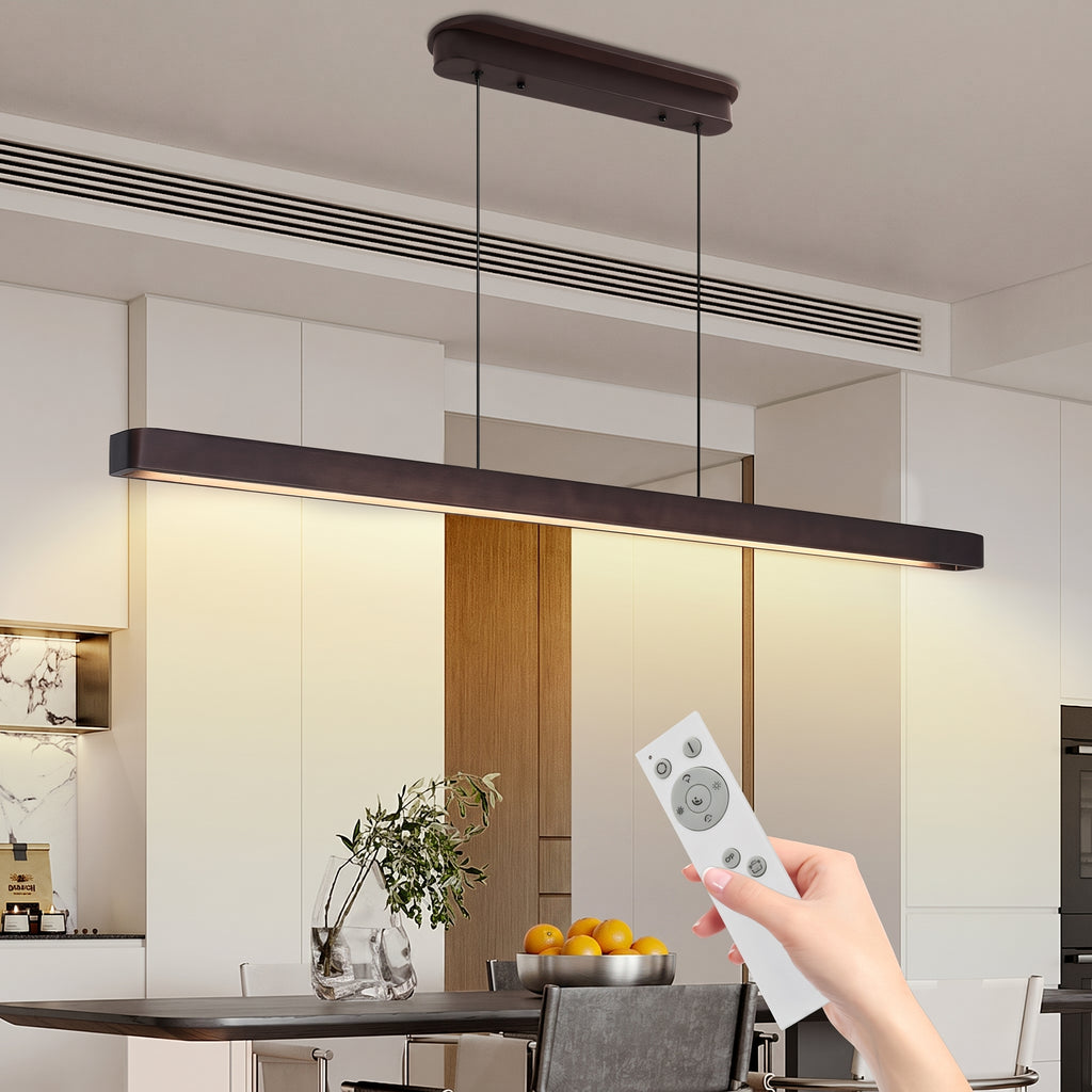 A modern linear LED pendant light with a dark wood-look frame hangs over a dining table in a bright kitchen or dining area. A hand holds a white remote control in the foreground, suggesting wireless control.