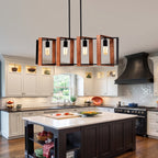 A modern kitchen island with a white countertop and dark wood cabinetry, illuminated by a rectangular pendant light with copper-toned side panels and black metal framing. The kitchen features white upper cabinets, a black range hood, and wood flooring.