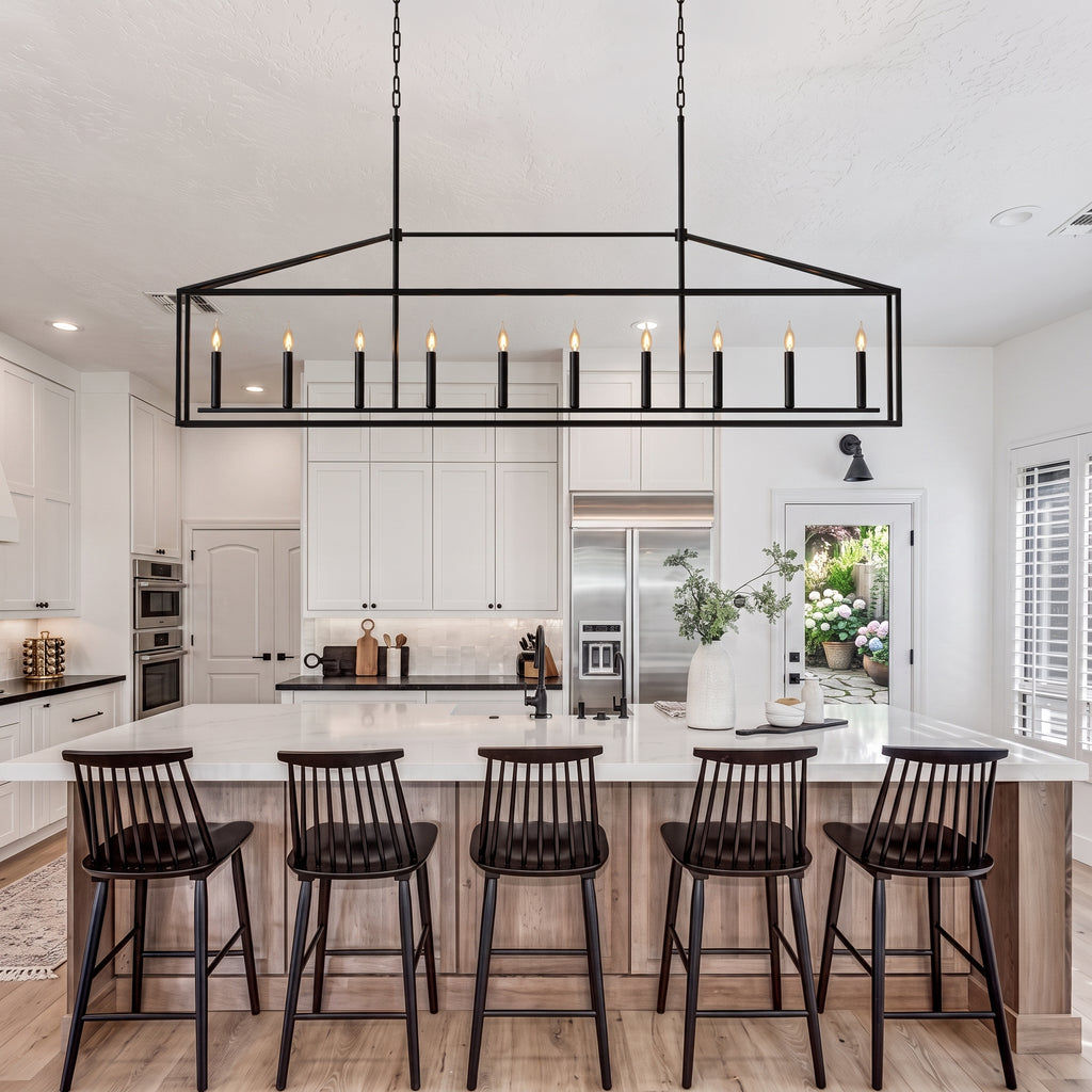 A modern kitchen island with five black spindle-back bar stools under a large rectangular black metal pendant light with candle-style bulbs. The kitchen features white cabinets, a white marble island, light wood flooring, and stainless steel appliances.