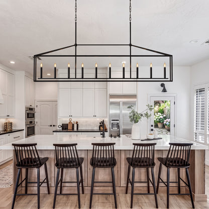 A modern kitchen island with five black spindle-back bar stools under a large rectangular black metal pendant light with candle-style bulbs. The kitchen features white cabinets, a white marble island, light wood flooring, and stainless steel appliances.