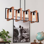 A modern 4-light linear pendant chandelier with a black metal frame, copper-toned wooden geometric brackets, and exposed Edison-style bulbs, shown hanging in a styled room setting with a framed photo, plant, and globe.