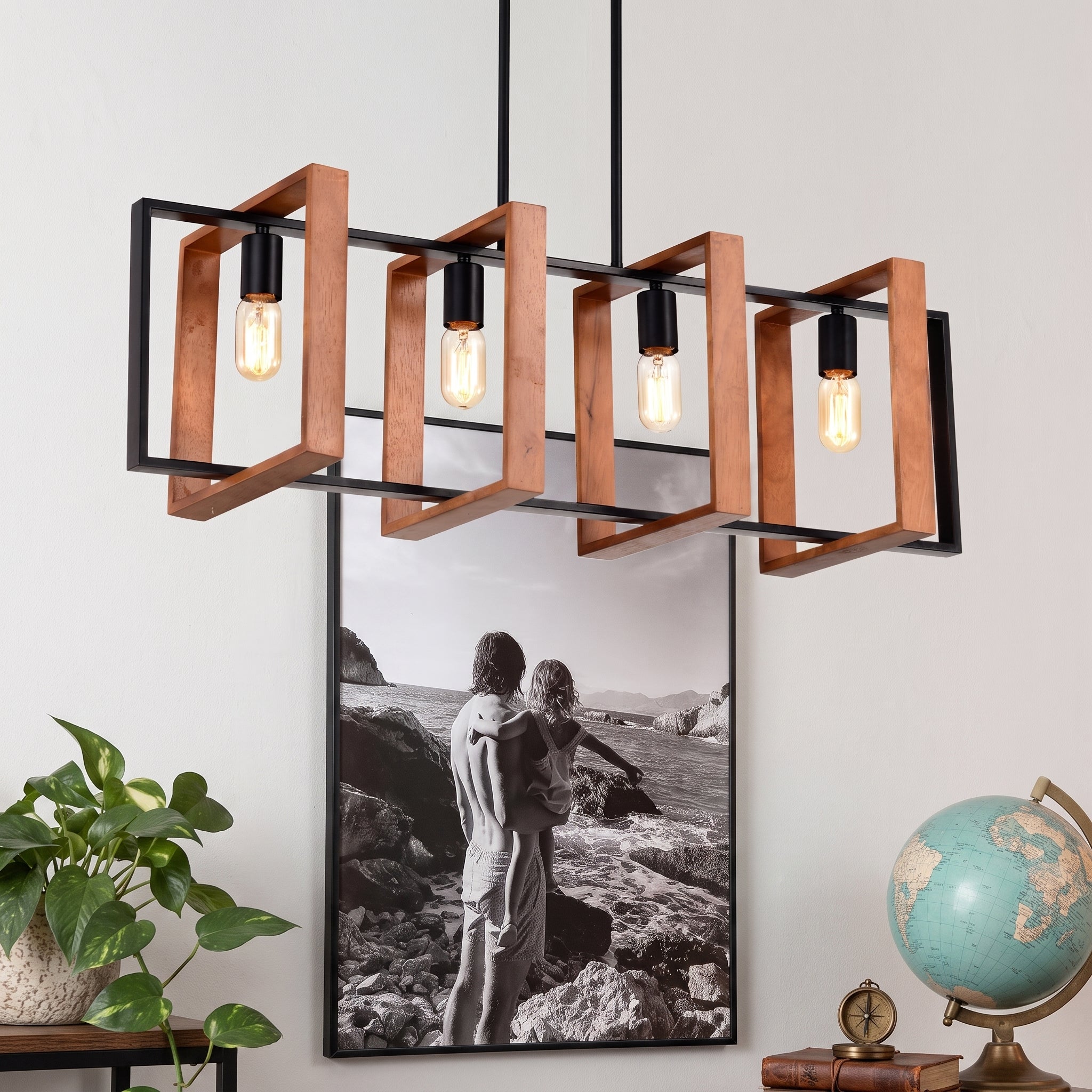 A modern 4-light linear pendant chandelier with a black metal frame, copper-toned wooden geometric brackets, and exposed Edison-style bulbs, shown hanging in a styled room setting with a framed photo, plant, and globe.