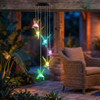 A solar-powered hummingbird wind chime hanging from a wooden porch at night, with glowing multicolor LED lights and a warm patio setting in the background.