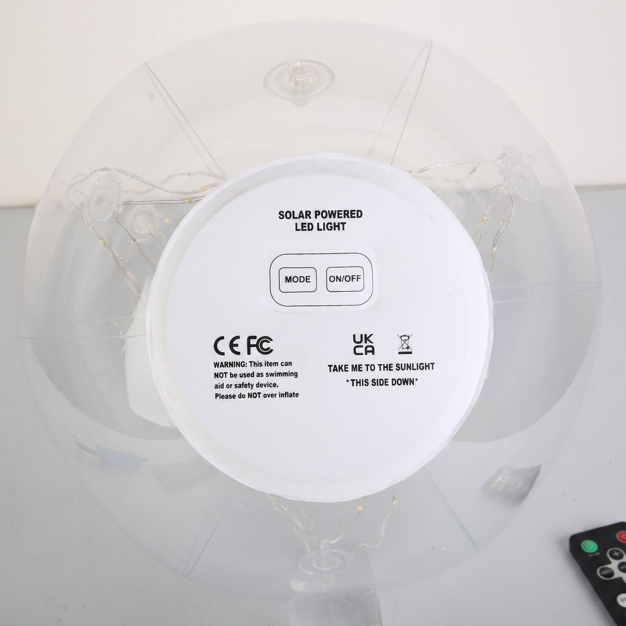 Top-down view of a solar-powered LED light on an inflatable ring, showing the white circular control panel with mode and on/off buttons, CE and UKCA certification marks, and a small remote control in the corner.