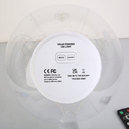 Top-down view of a solar-powered LED light on an inflatable ring, showing the white circular control panel with mode and on/off buttons, CE and UKCA certification marks, and a small remote control in the corner.