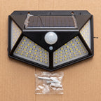 Black solar-powered outdoor wall light with an integrated motion sensor, shown on a brown cardboard background with mounting screws in a small plastic bag below.