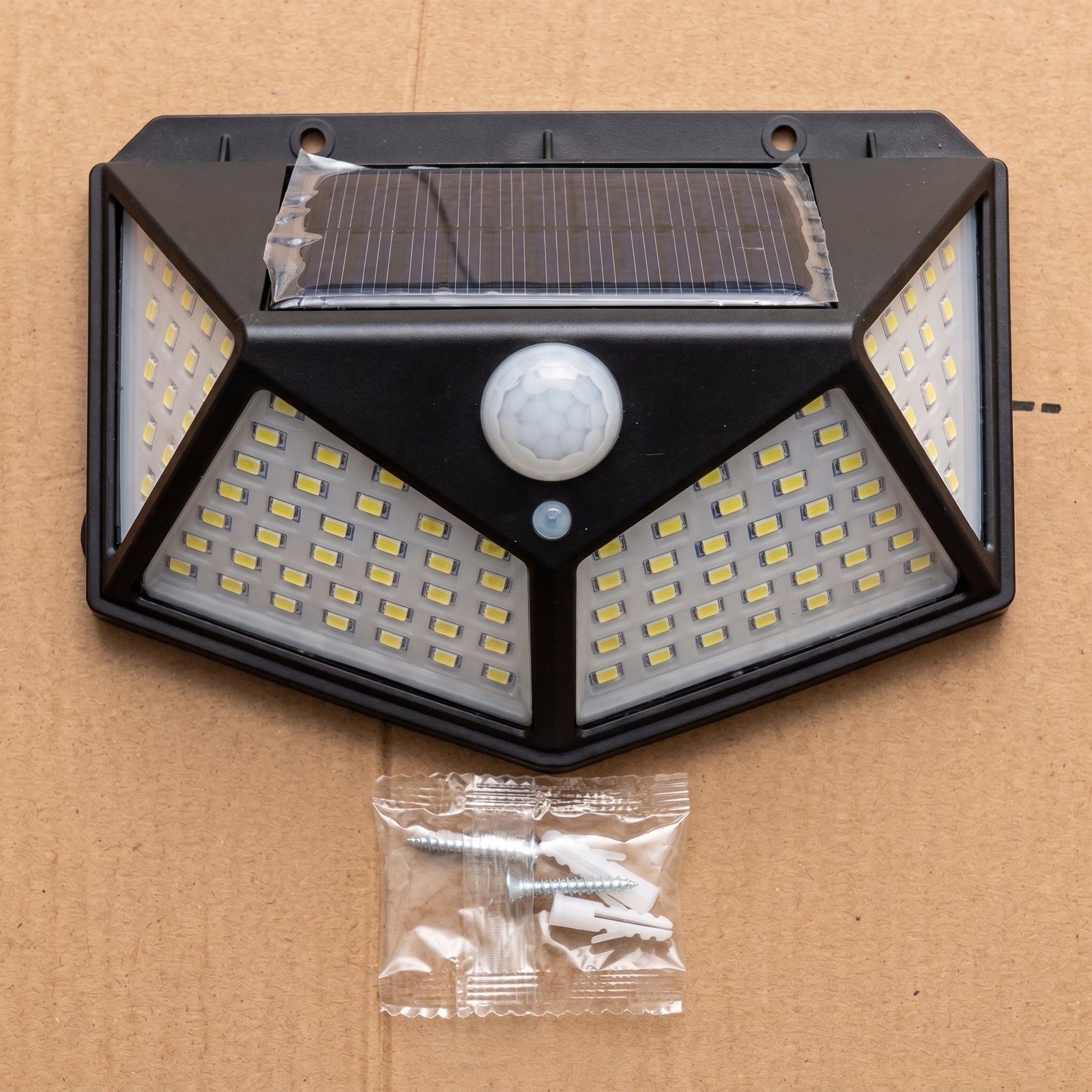 Black solar-powered outdoor wall light with an integrated motion sensor, shown on a brown cardboard background with mounting screws in a small plastic bag below.